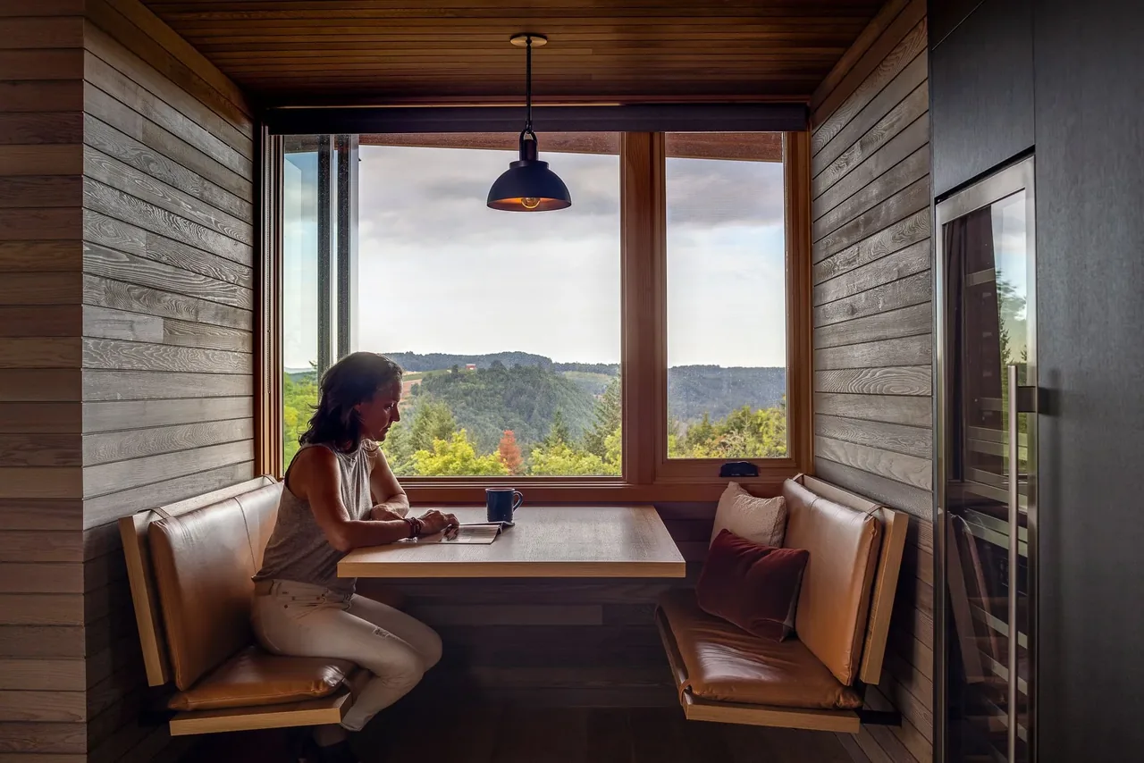 Built-in leather bench seating and wooden table occupy a dining nook with a panoramic forest view at Lone Feather Residence.