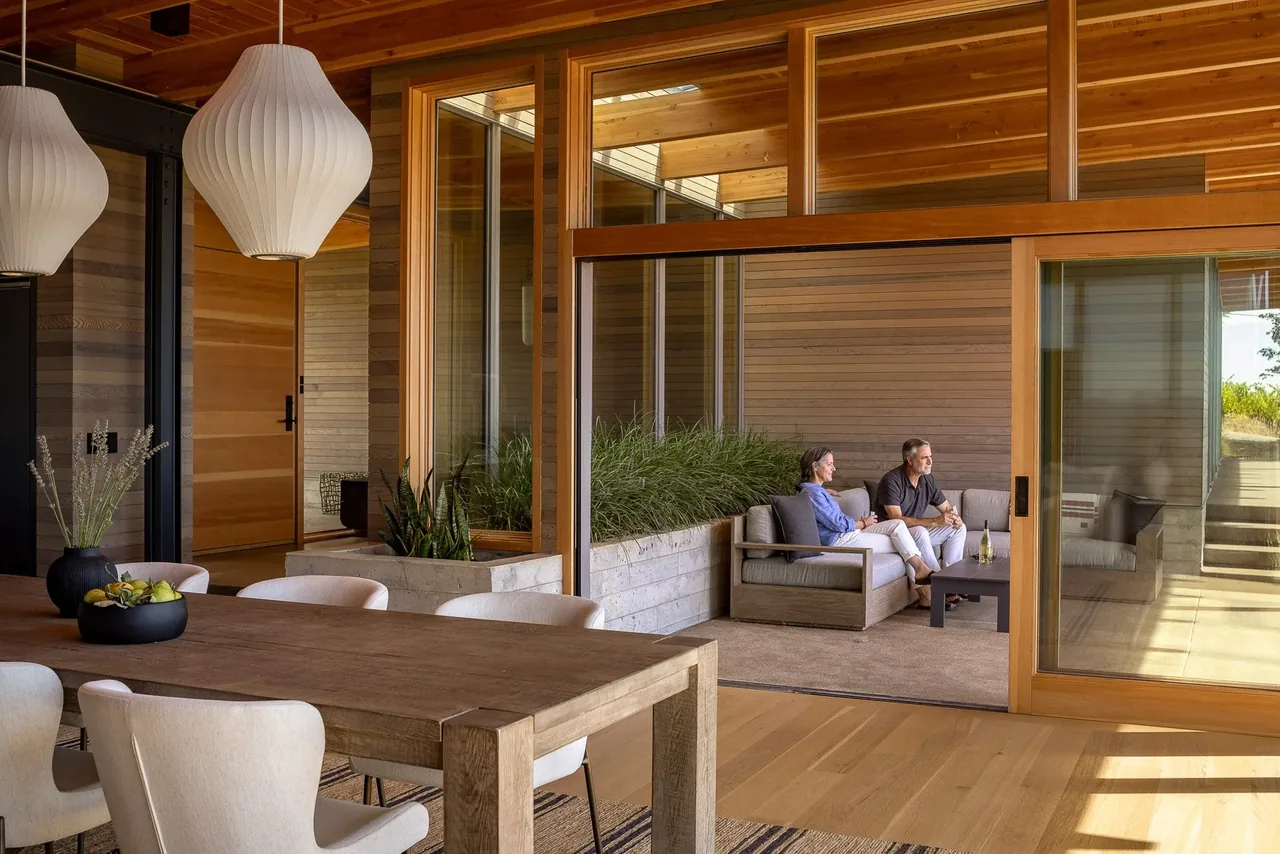An open dining room with wood ceiling beams seamlessly connects to an outdoor living space with concrete planters and large glass doors.