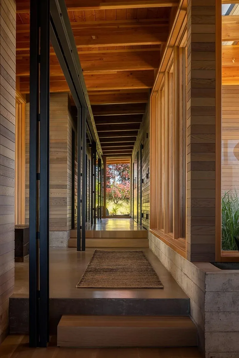 The hallway features wood paneling on walls and ceiling, concrete base, and steel framed glass doors revealing outdoor greenery.