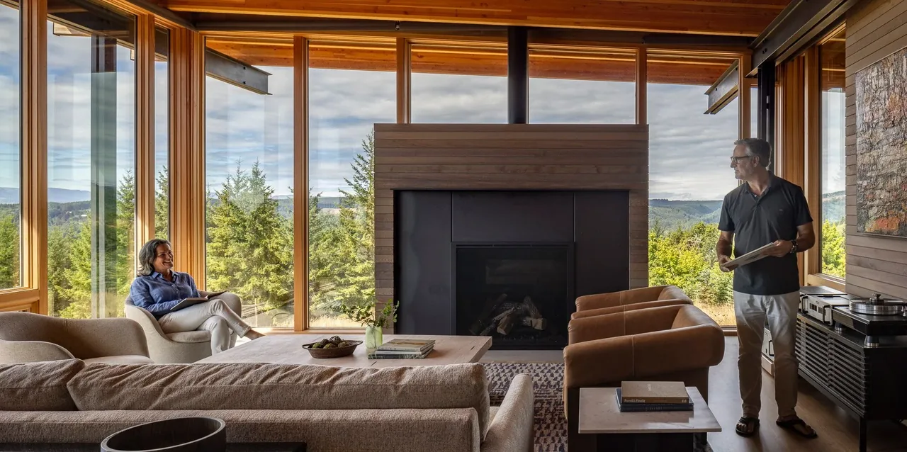 The Lone Feather Residence living room features floor-to-ceiling windows and a wood-paneled fireplace framed by serene forest views.