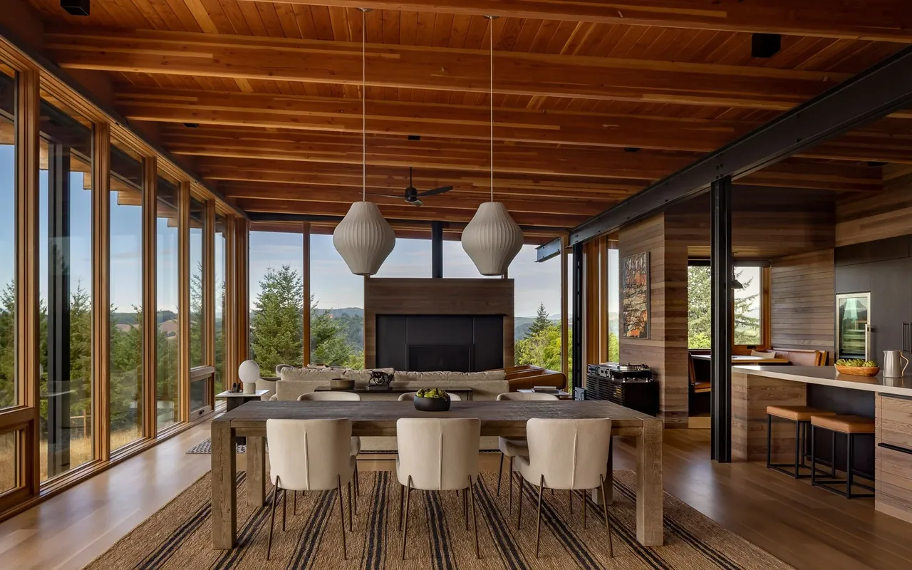 Open living and dining space with wood finishes and expansive glass walls overlooking a forested landscape.