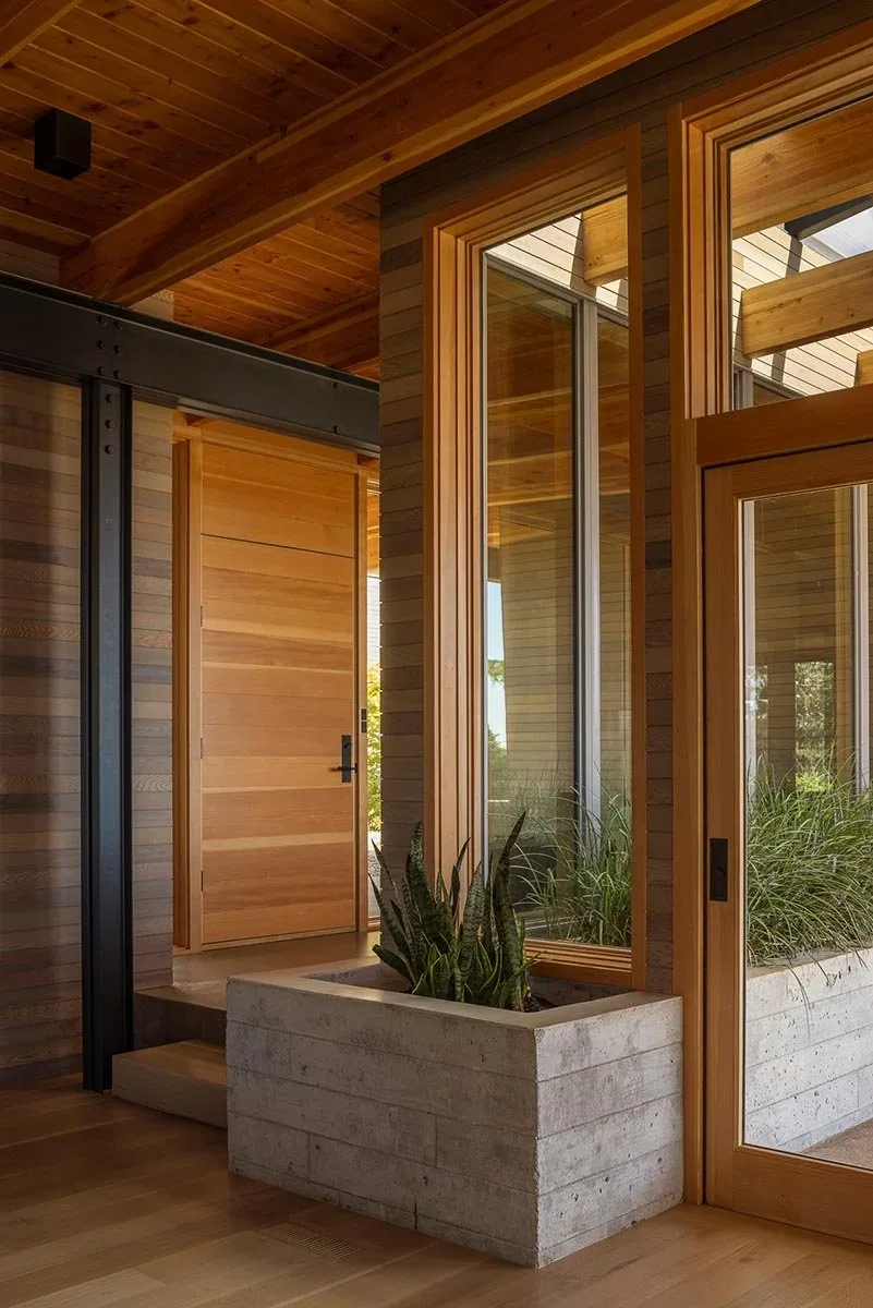 The Lone Feather Residence entry features wood ceiling beams, large wood-framed windows, and a concrete planter box with greenery.