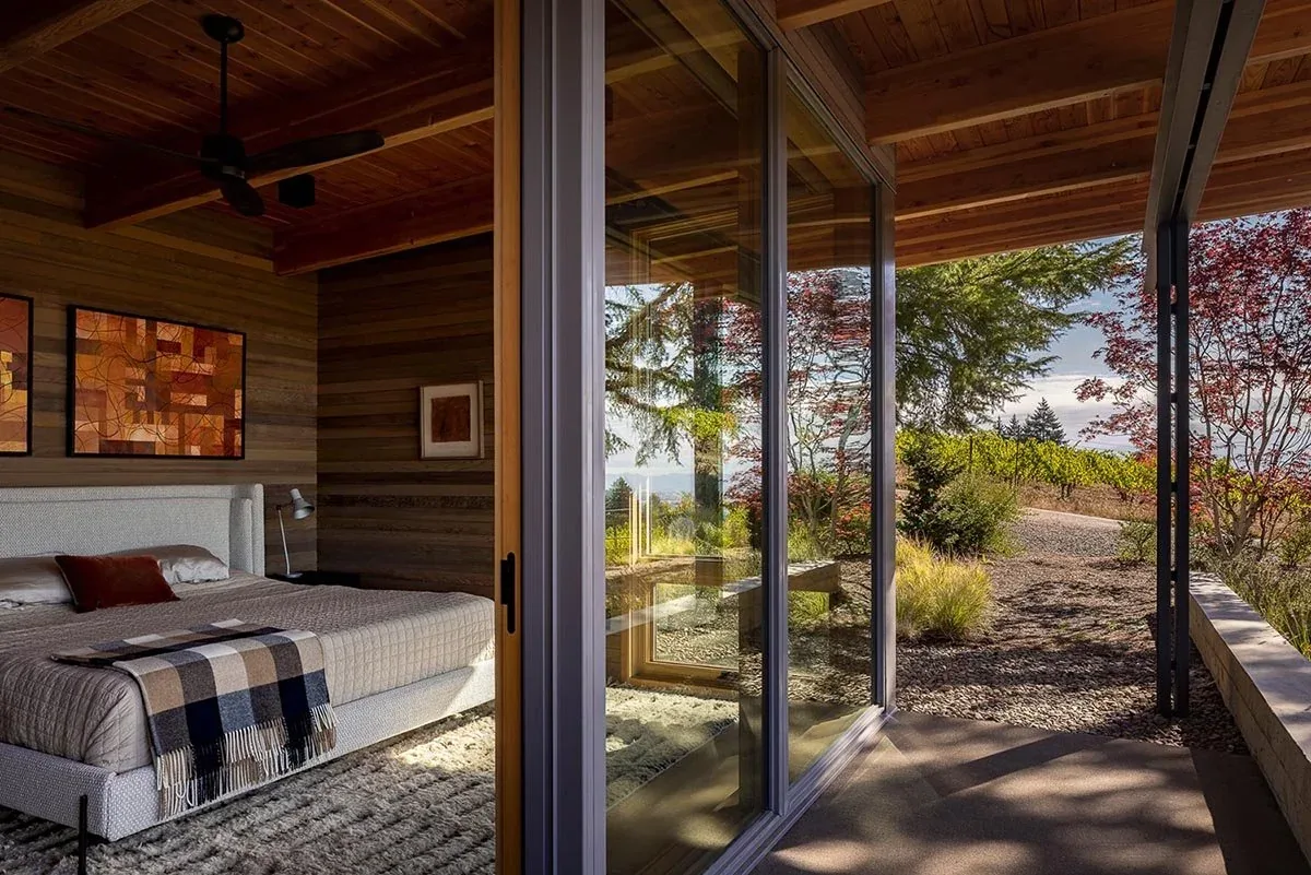 The Lone Feather Residence bedroom shows wood walls and ceiling beams with a sliding glass door leading to a patio with a vineyard view.