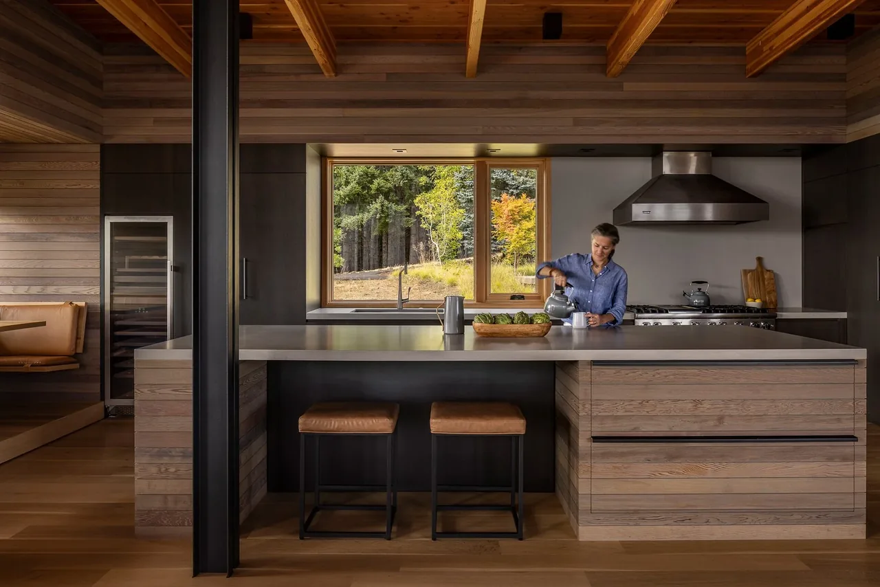 Contemporary kitchen featuring wood beams, leather cushioned stools, and a panoramic forest view through a large window.