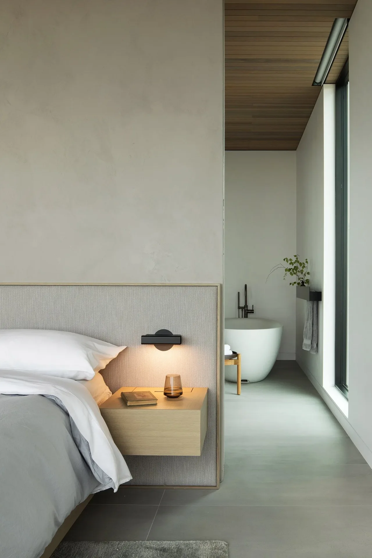 Minimal bedroom with a floating wooden nightstand and adjacent bathroom featuring a freestanding tub and black fixtures.