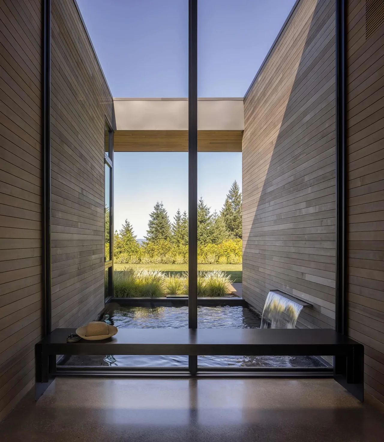 A bench inside faces a water feature with a waterfall and landscaped greenery beyond the wood-paneled walls.