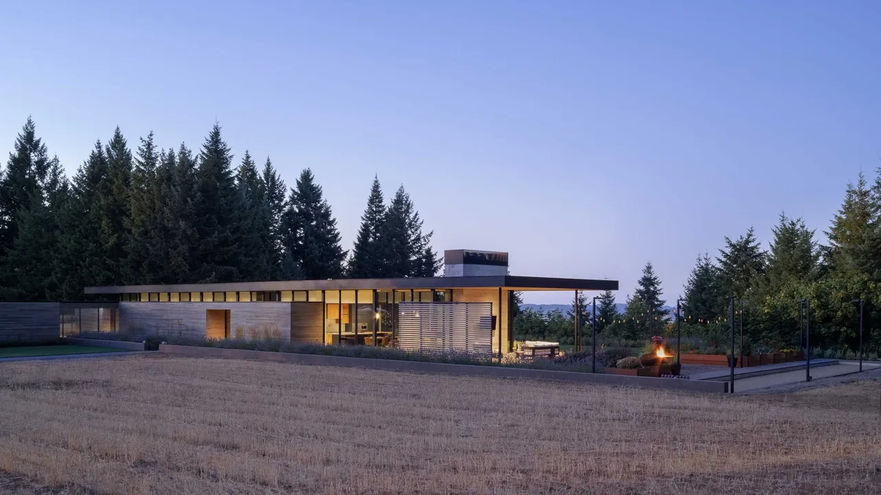 Modern flat-roofed residence with expansive glazing and outdoor seating area at dusk, set against a forested landscape.