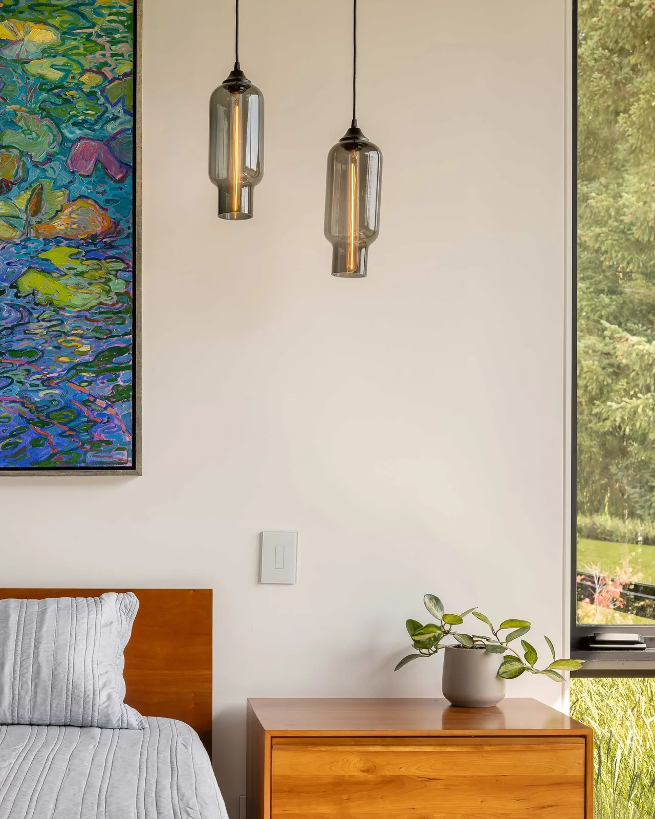 Wood nightstand next to a bed with gray bedding, two glass pendant lights, and a large window revealing greenery outside.