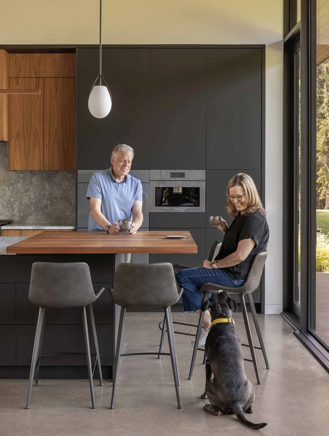 Modern kitchen features gray matte cabinets, wood breakfast bar, and polished concrete floor near corner glass windows.