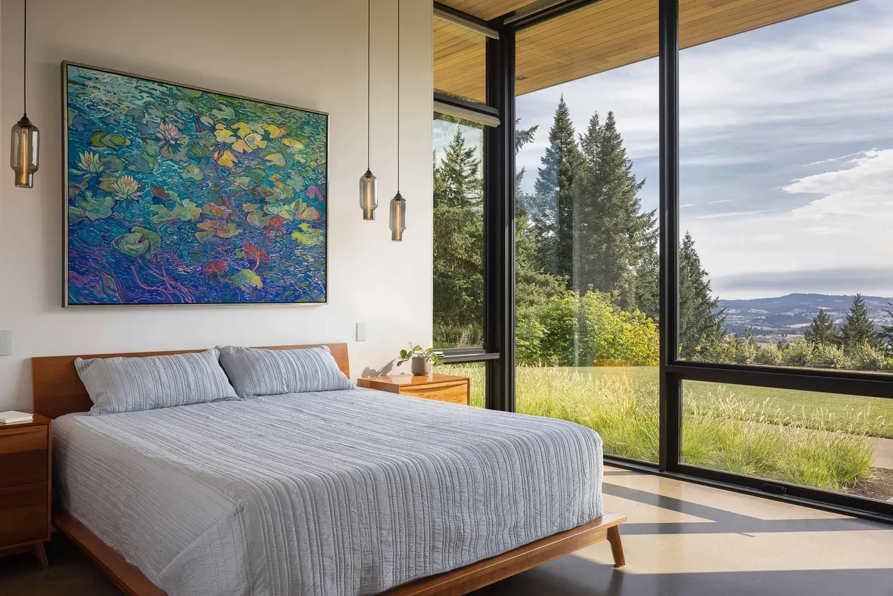 The bedroom features large black-framed floor-to-ceiling windows with a view of trees and hills.