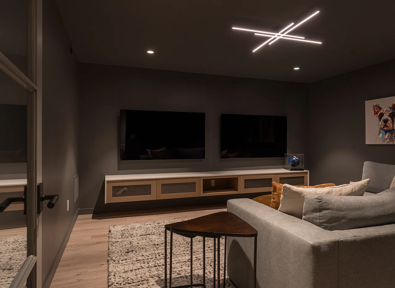 Media room with dual flat screen TVs mounted above a floating console and a gray sofa under linear ceiling lights.