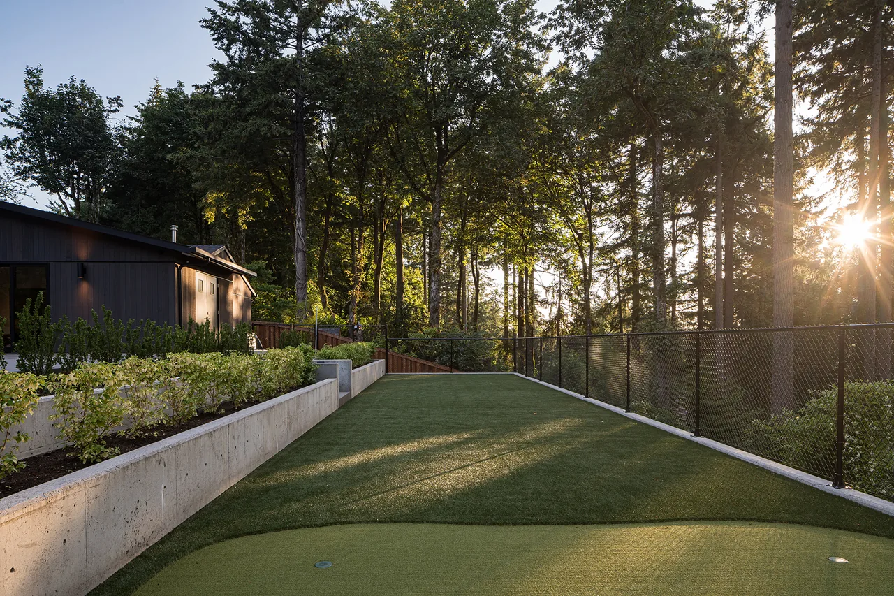 A backyard putting green beside a modern house with black siding and a retaining wall under tall trees.