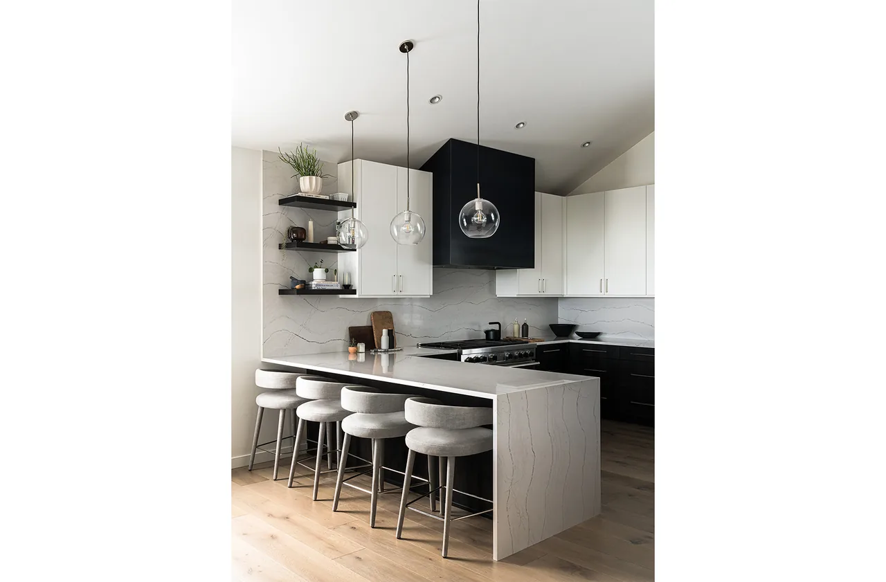 Fernwood House kitchen featuring a marble island with seating, suspended glass globe pendants, and a black and white cabinetry contrast.