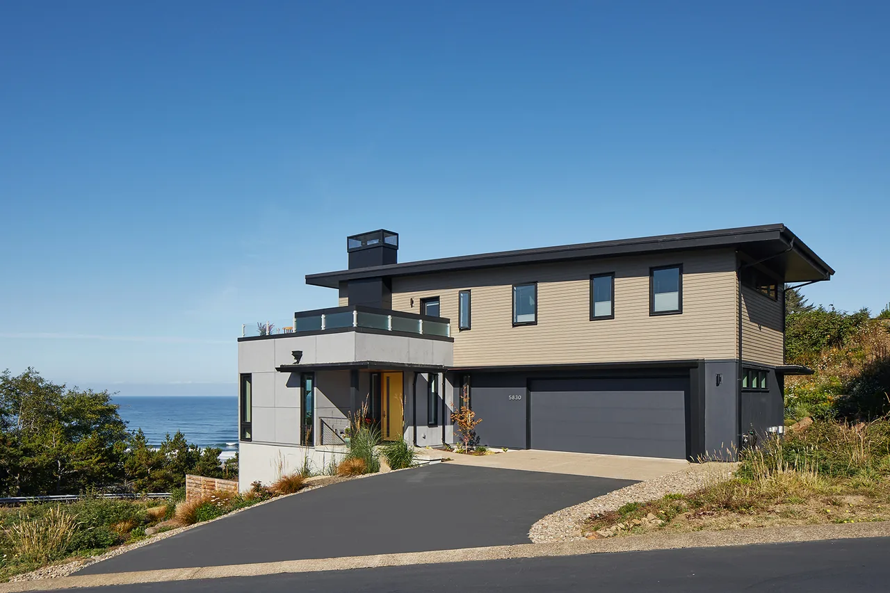 Sahhali Shores Shelter is a modern two-story house featuring beige wood siding, black window frames, and a flat roof with ocean views.
