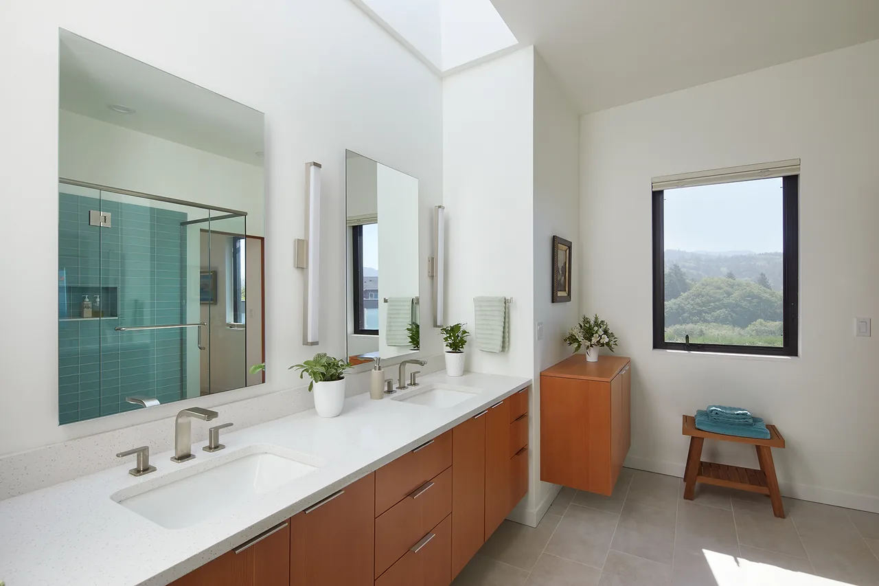 Bathroom with dual sink vanity, wooden cabinets, skylight, and glass shower with teal tile visible.