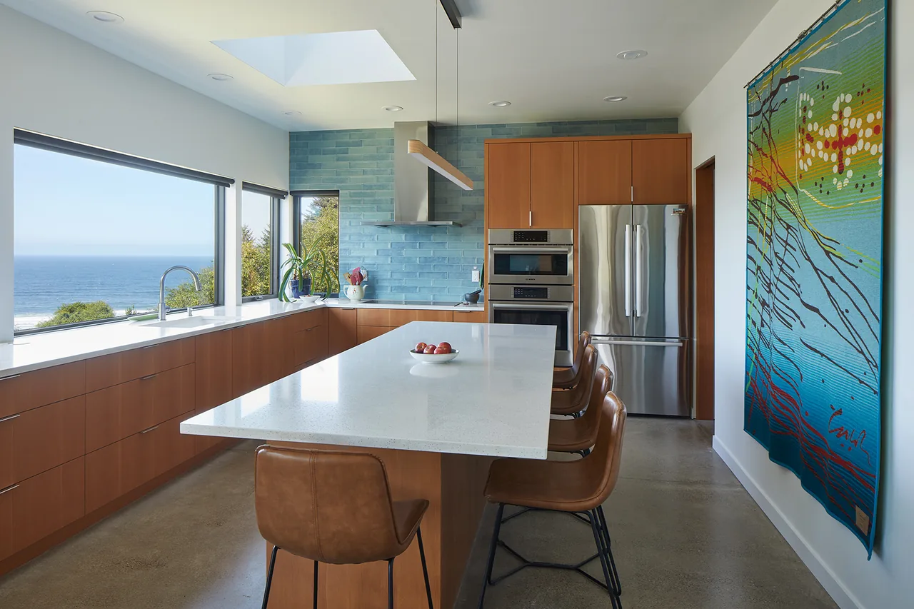 The kitchen in Sahhali Shores Shelter features wood cabinetry, a blue tile backsplash, and large windows overlooking the ocean.