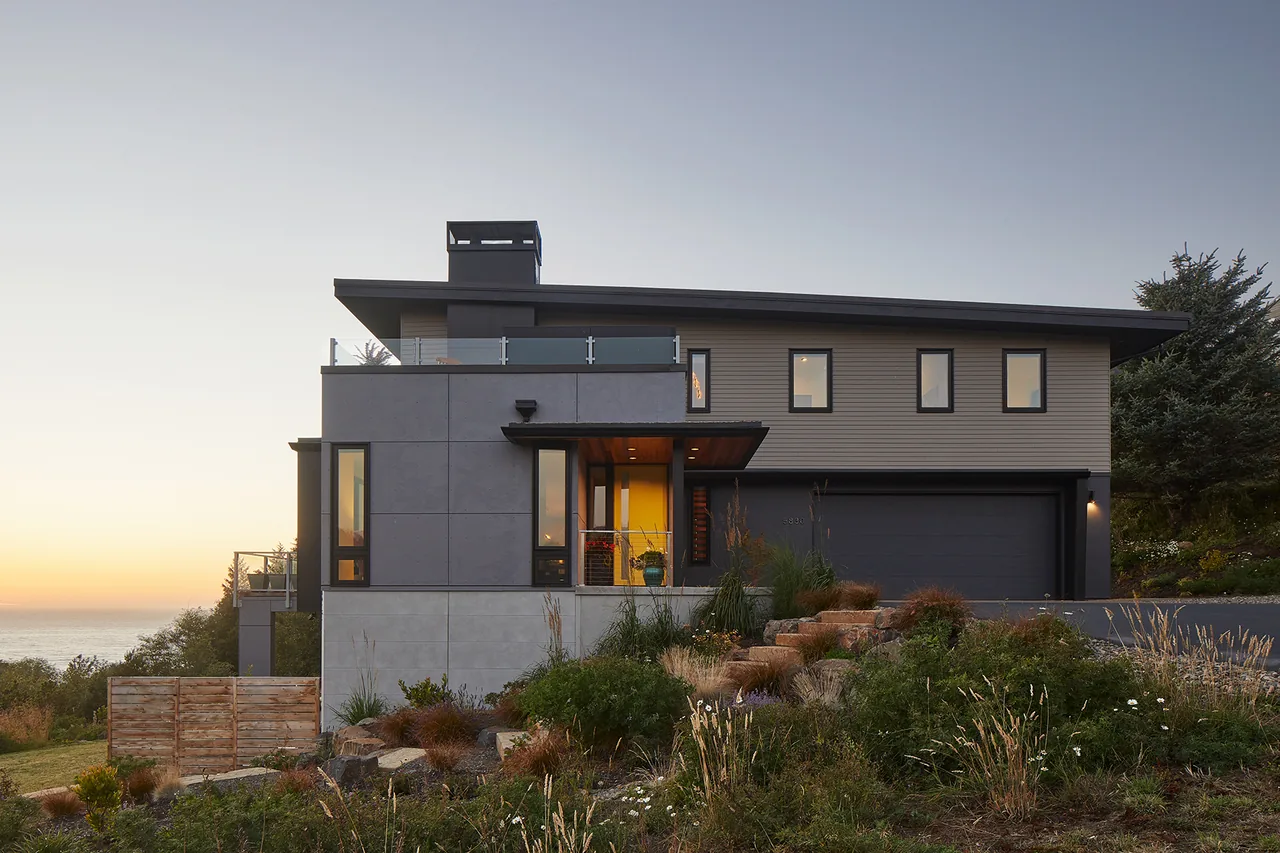Sahhali Shores Shelter is a modern multi-story house featuring black window frames and a driveway, overlooking the ocean at sunset.