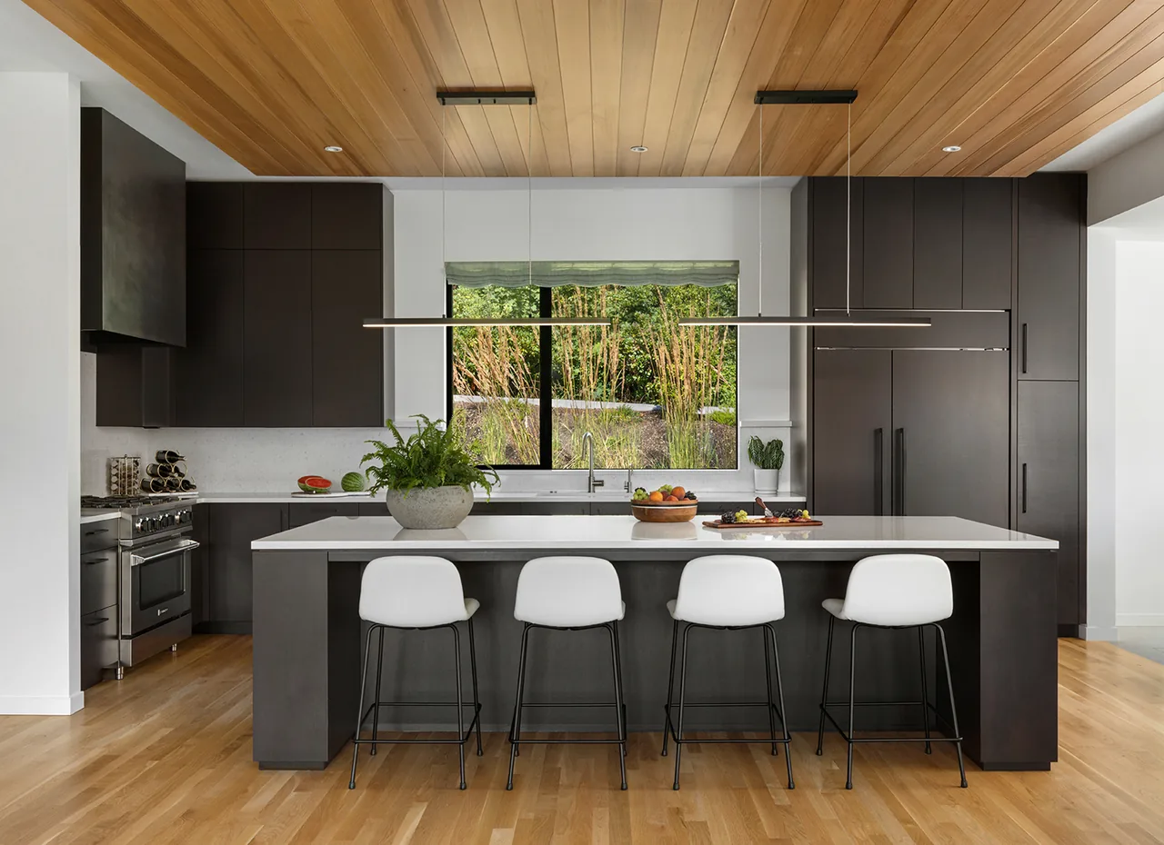 The Hilltop House kitchen by Giulietti Schouten Weber Architects features dark cabinetry and a white island under a wood-paneled ceiling.