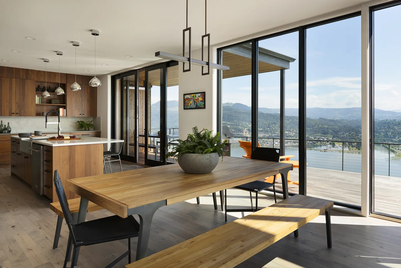 View of the dining table and kitchen with wood finishes and large windows showing a river landscape.