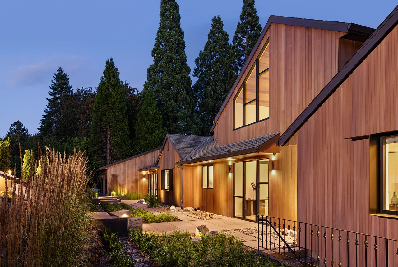 Hilltop House exterior shows wood siding, large windows, and pathway lighting with a gravel garden bed and natural landscaping.