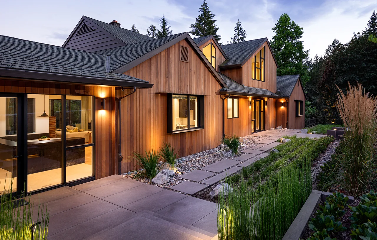 Hilltop House by Giulietti Schouten Weber Architects features wood siding, a stone pathway, and dusk lighting revealing interior living spaces.