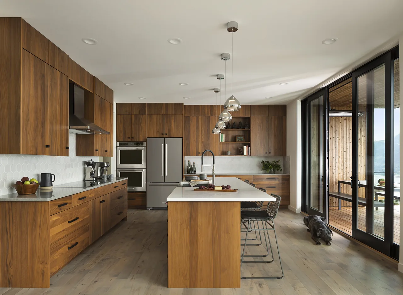 Hilltop House kitchen features walnut cabinets, a large island with pendant lights, and sliding glass doors that open to a deck with a forest and river view.