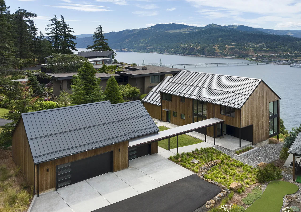 Hilltop House features standing seam metal roofs and vertical wood siding facing a river and hills.