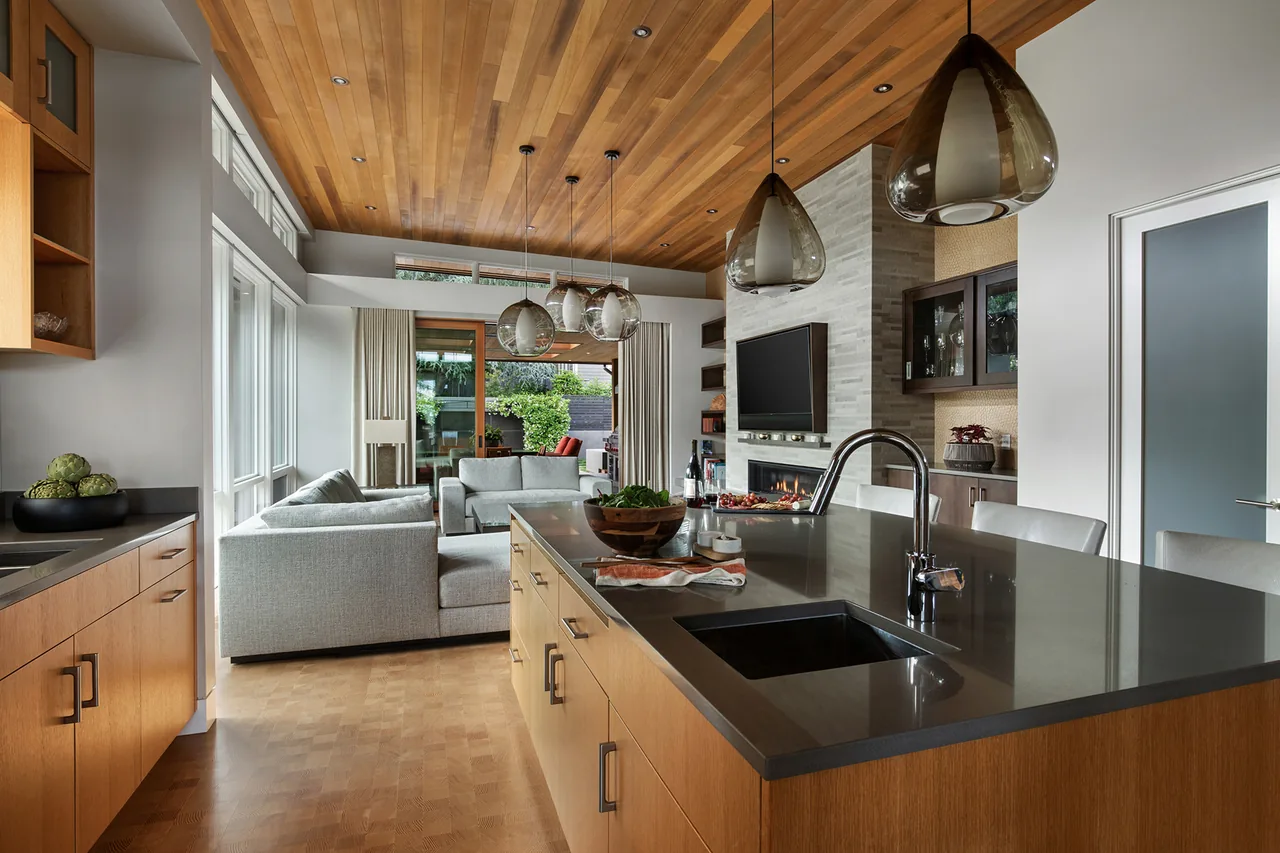 The Highlands Loop Residence features a wood ceiling living room and kitchen with large windows, a fireplace, and pendant lighting by Giulietti Schouten Weber Architects.
