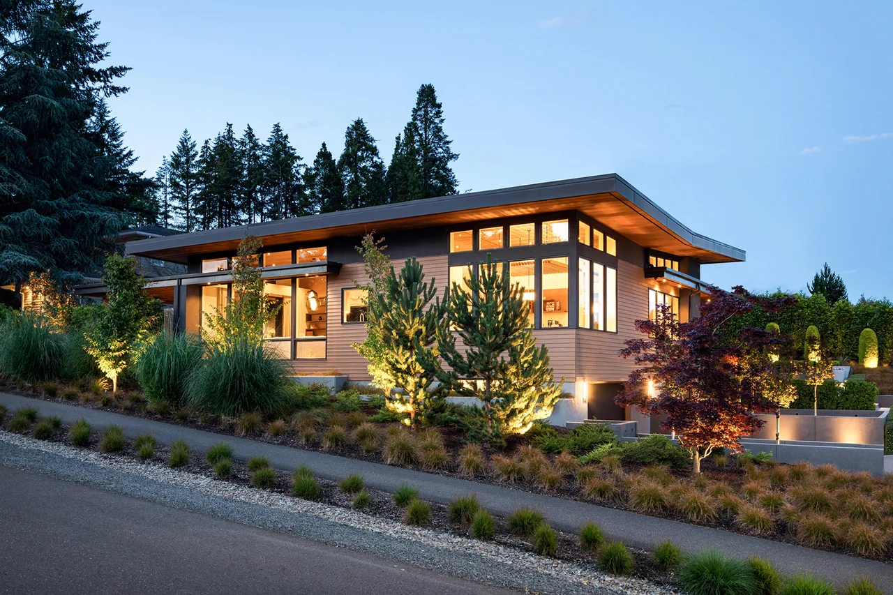 Exterior view of a modern house with a cantilevered flat roof and large corner windows, surrounded by landscaping at dusk.