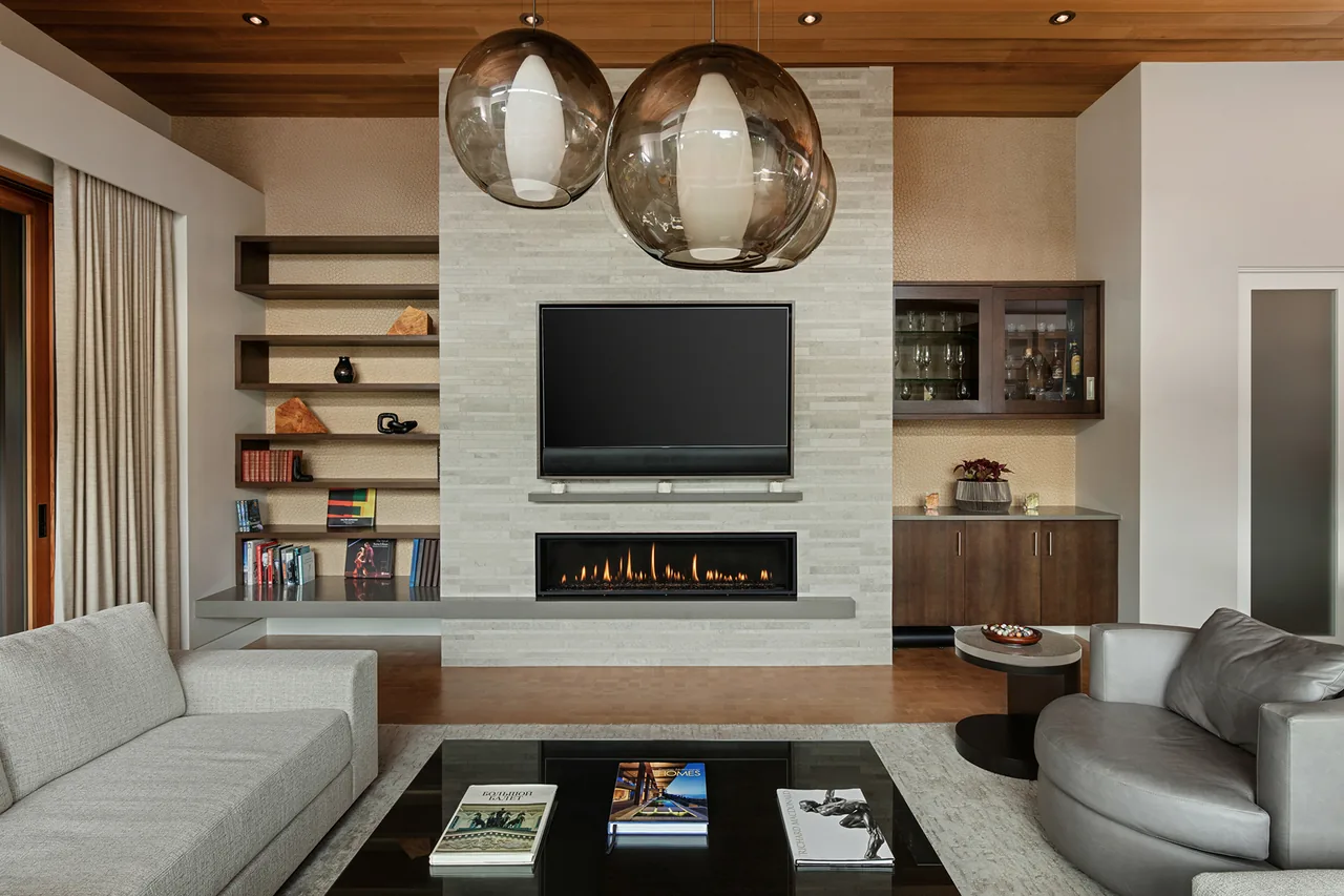 Living room features a tile fireplace wall with a mounted TV, floating shelves, a gray sofa, and a round leather chair under wood ceiling.