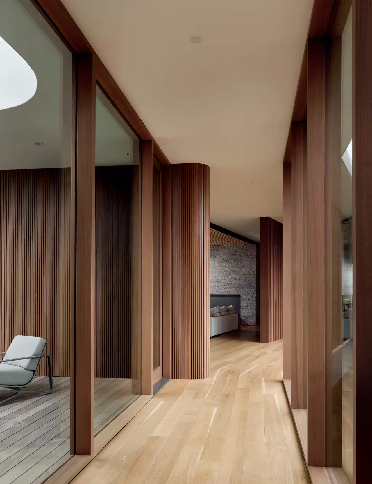 Hallway with floor-to-ceiling glass windows and curved wood-paneled wall leading to a living space with a stone fireplace.