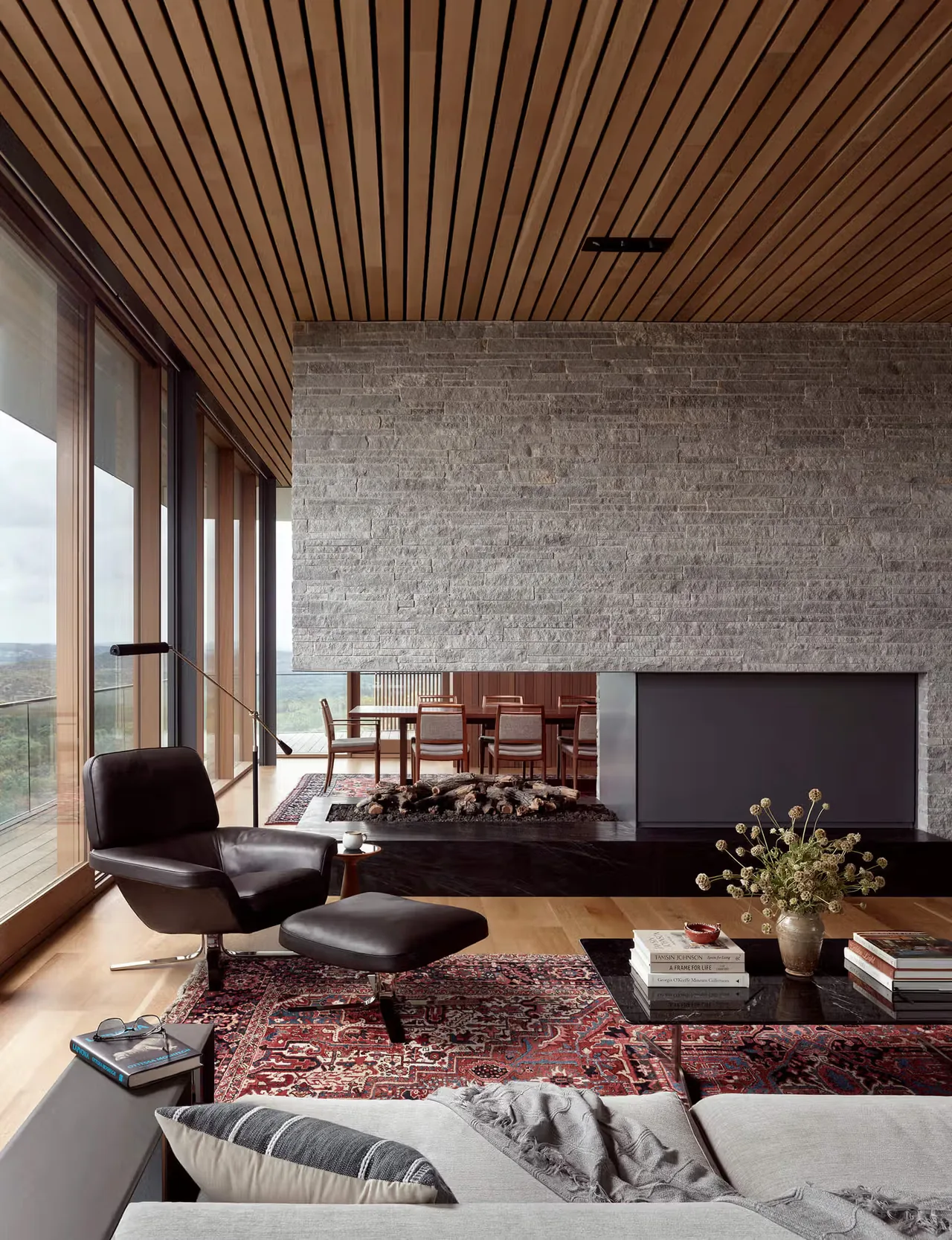 Living area features a large stone fireplace wall, floor-to-ceiling glazing, and wood panel ceiling.