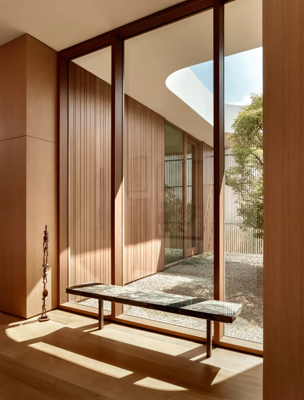 Natural light fills a hallway with wood-paneled walls and floor-to-ceiling glazing overlooking a courtyard with gravel and trees.