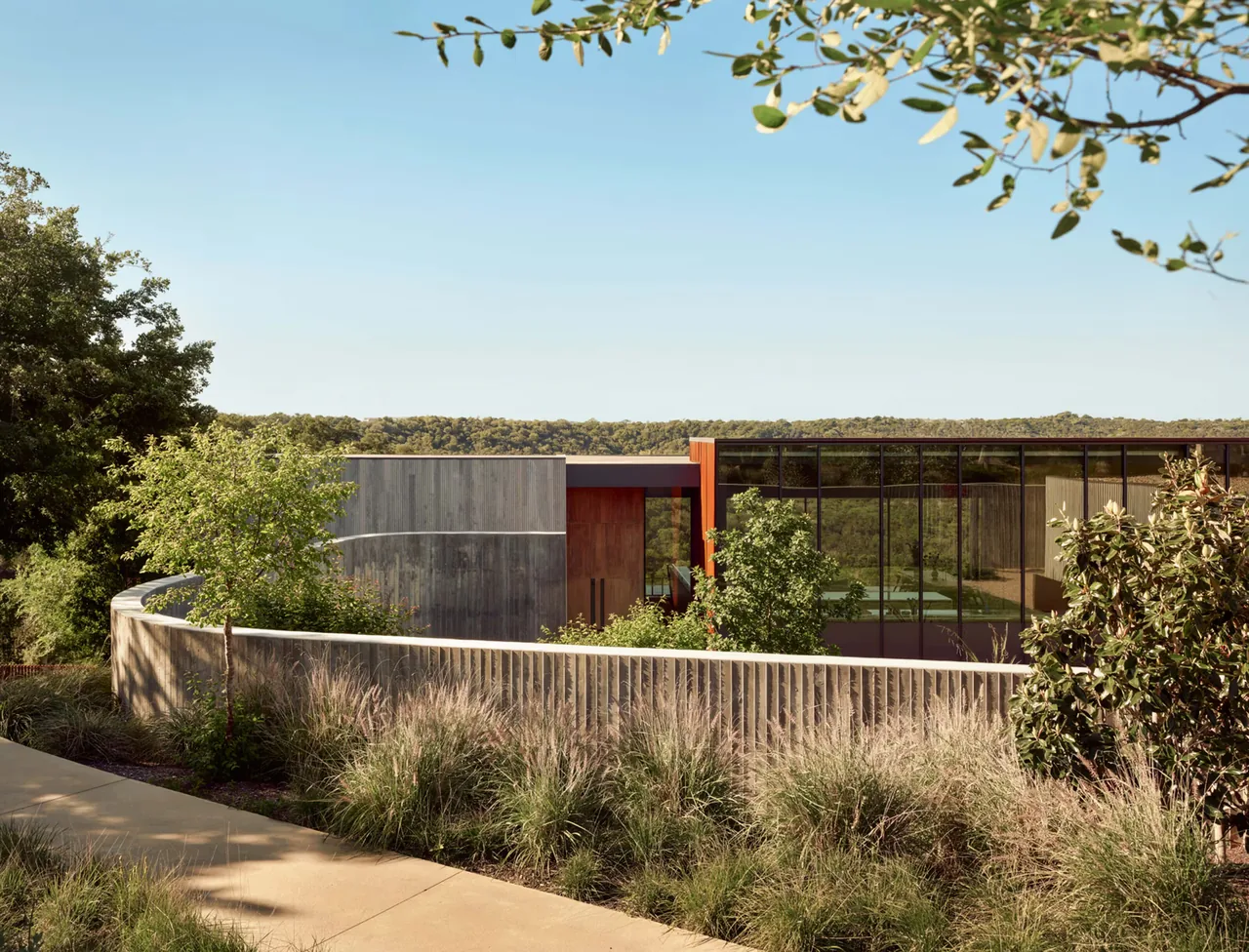 Barton Hills Residence features a flat roof, large glass facade with black window frames, and a wood-paneled entry, set behind a concrete perimeter wall.