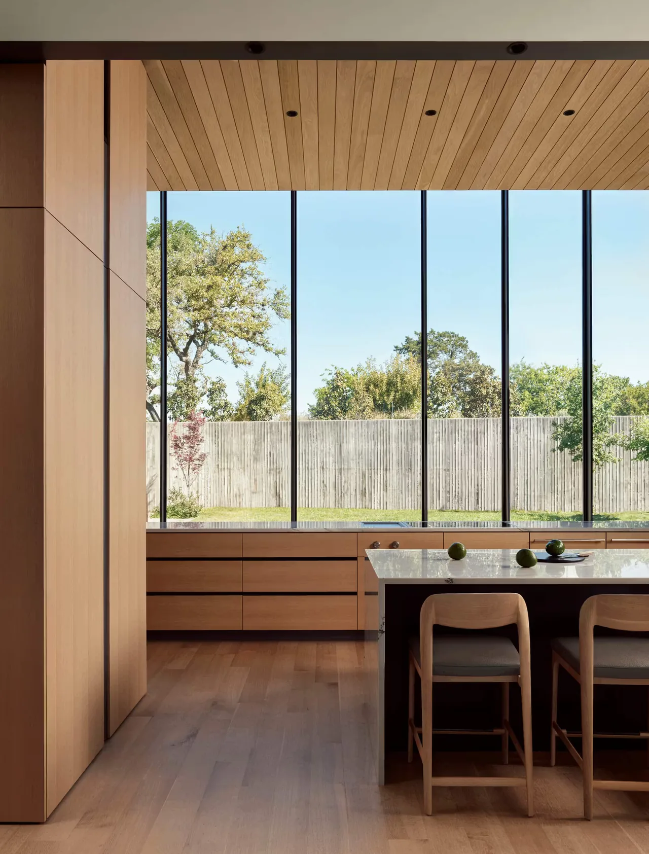 Kitchen space with wood-paneled cabinets and a large glass facade overlooking a yard with a wooden fence.