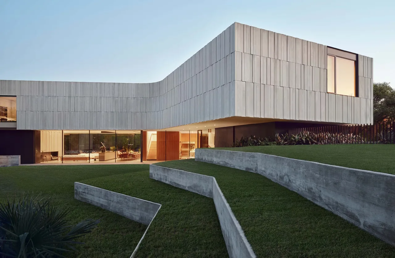 The Highland Park Residence features a wooden-clad upper floor over a glass-walled lower level with terraced concrete retaining walls in the front yard.