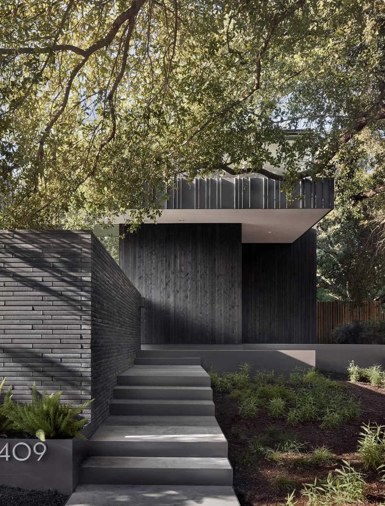 Modern residence showcasing black vertical wood siding with concrete steps and lush landscaping in front.