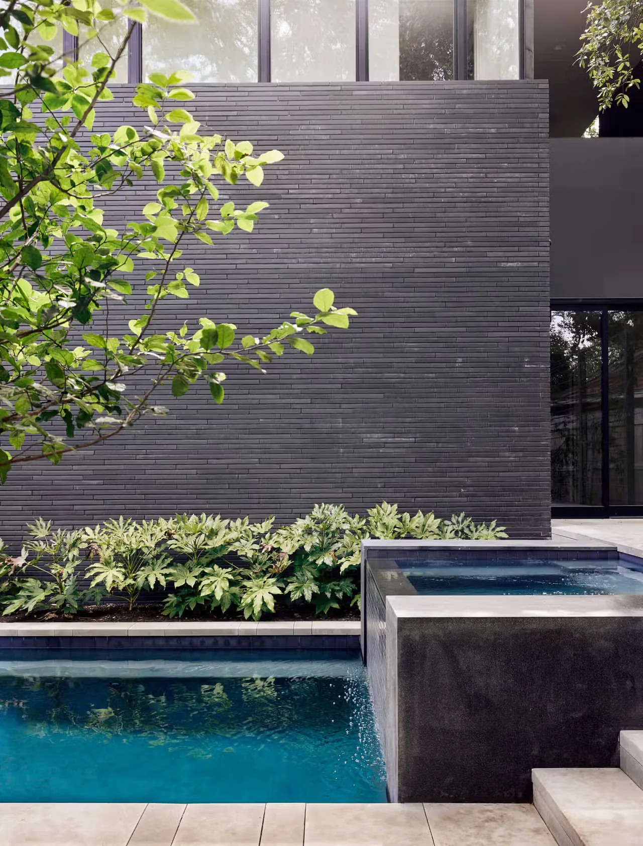 The Tarrytown Residence features a sleek black brick exterior wall adjacent to a pool with concrete steps and surrounding greenery.