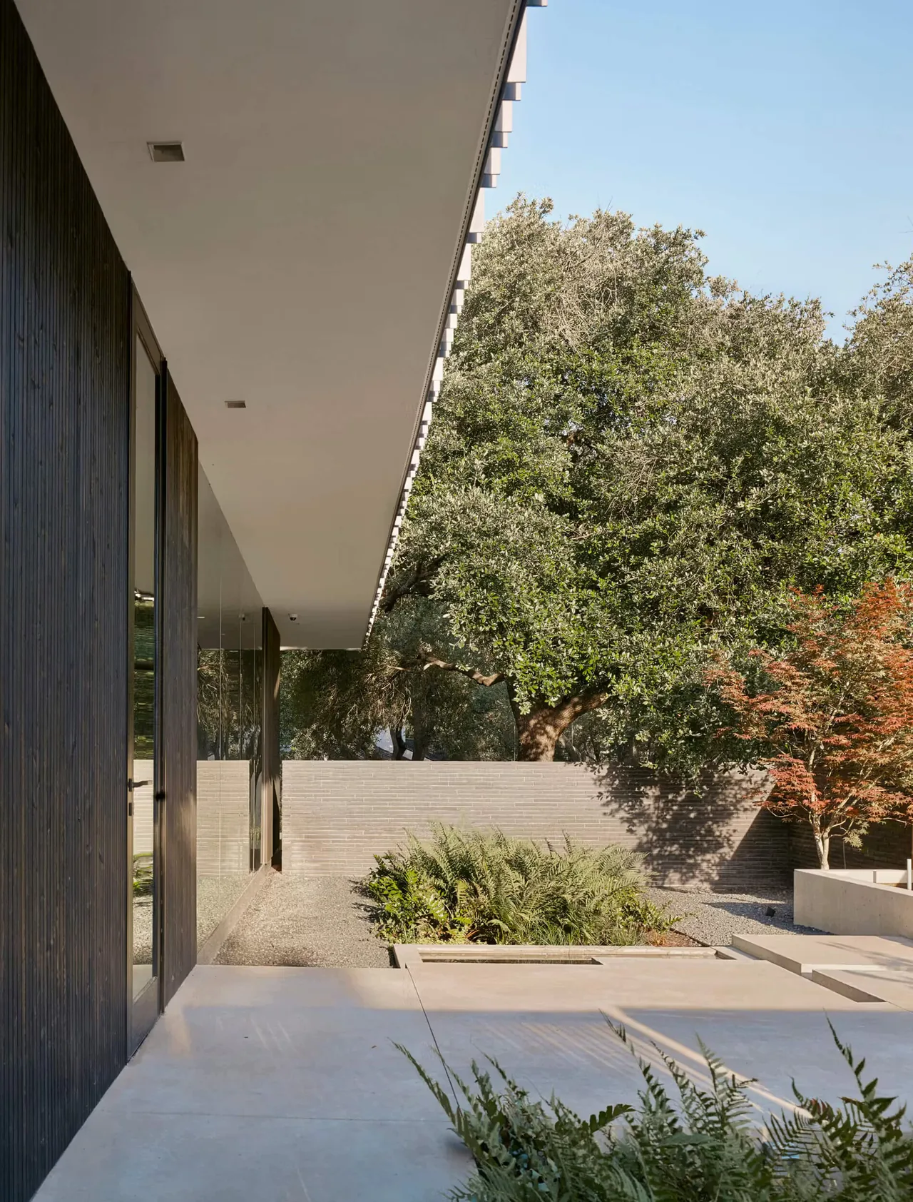Tarrytown Residence features a modern patio area with black wood siding and a well-landscaped garden with trees and shrubs.