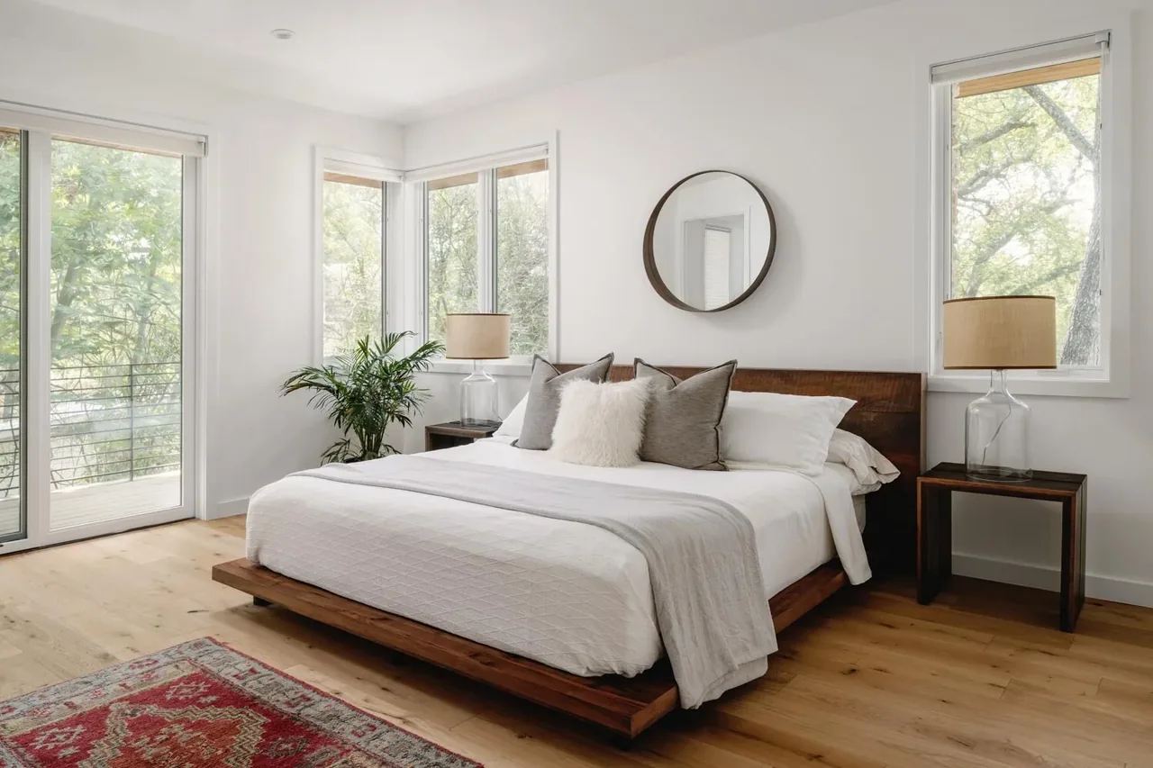 A minimalist bedroom with a wooden platform bed, large windows, neutral bedding, and natural light.