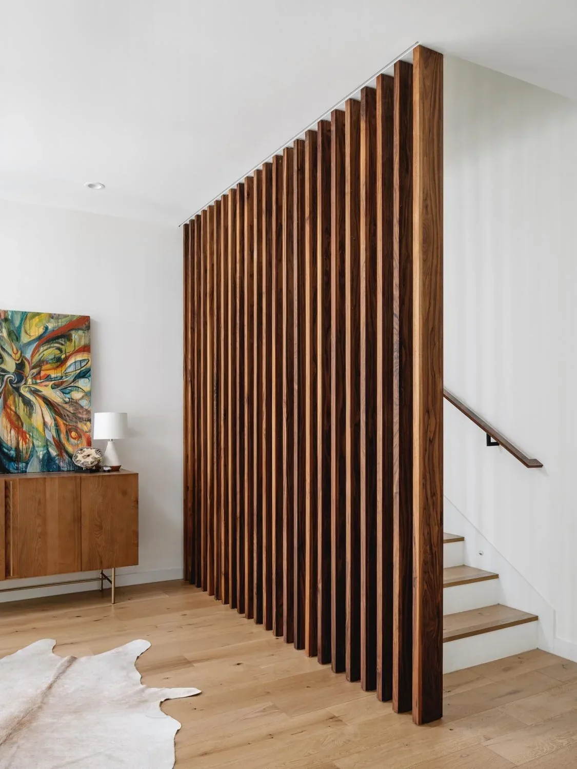 Vertical wood slats divide a living area and a staircase with wood flooring and a minimal handrail.