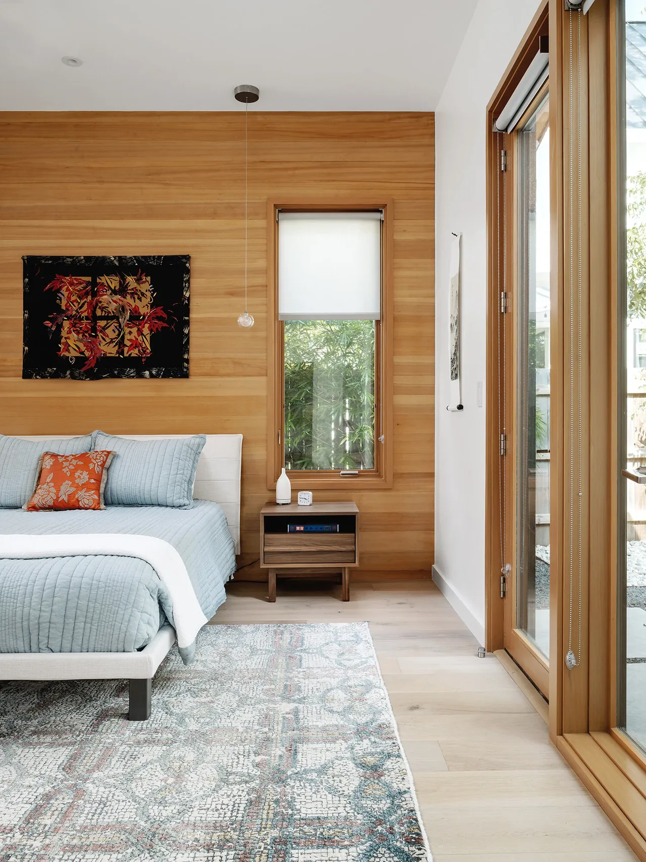 Bedroom in the Ramsey residence featuring wood paneled walls, a window with roller shade, and blue quilted bedding.