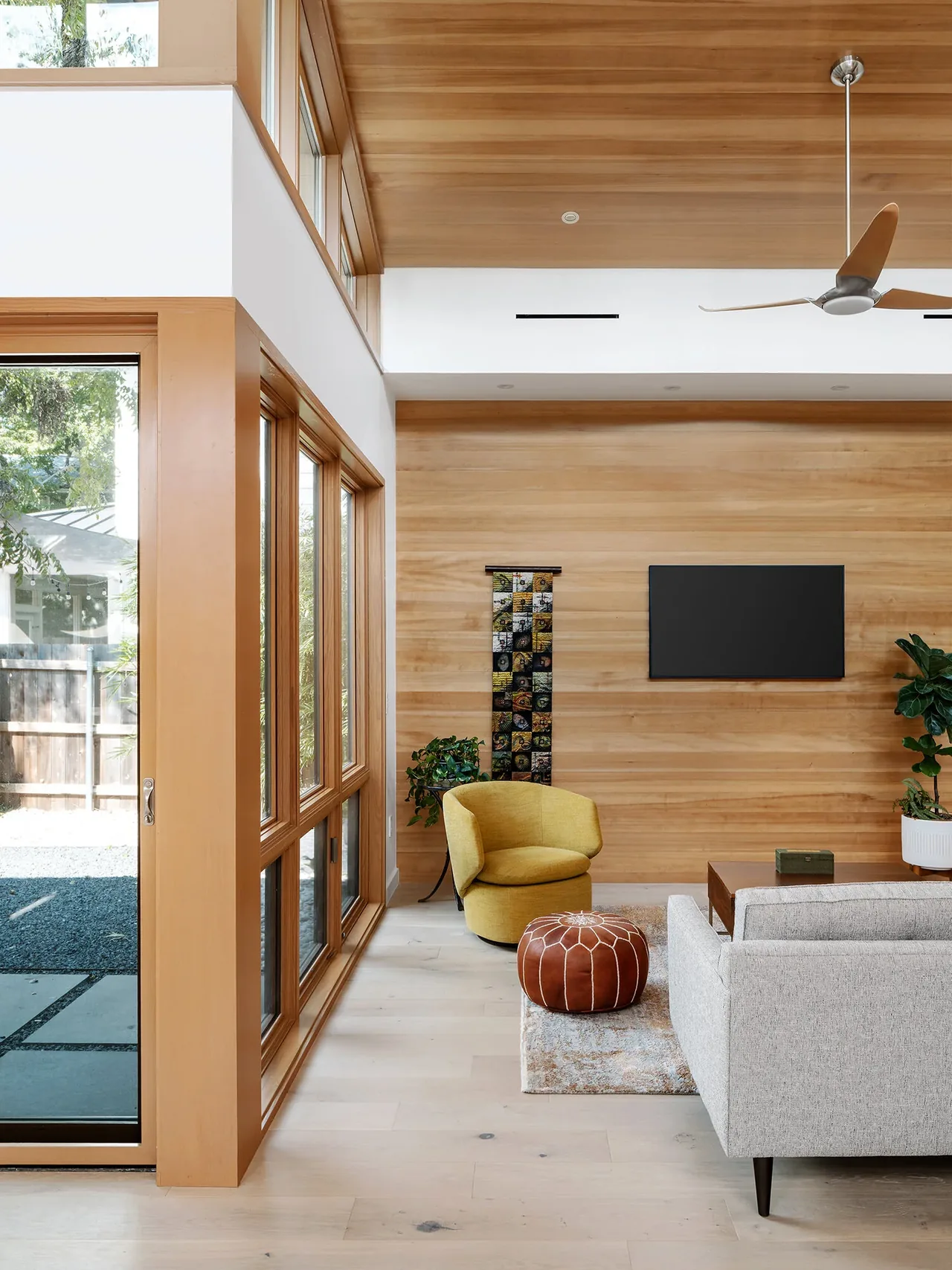 Living room of the Ramsey Residence features a wood-paneled wall, large windows, and modern furnishings including a gray sofa and yellow armchair.