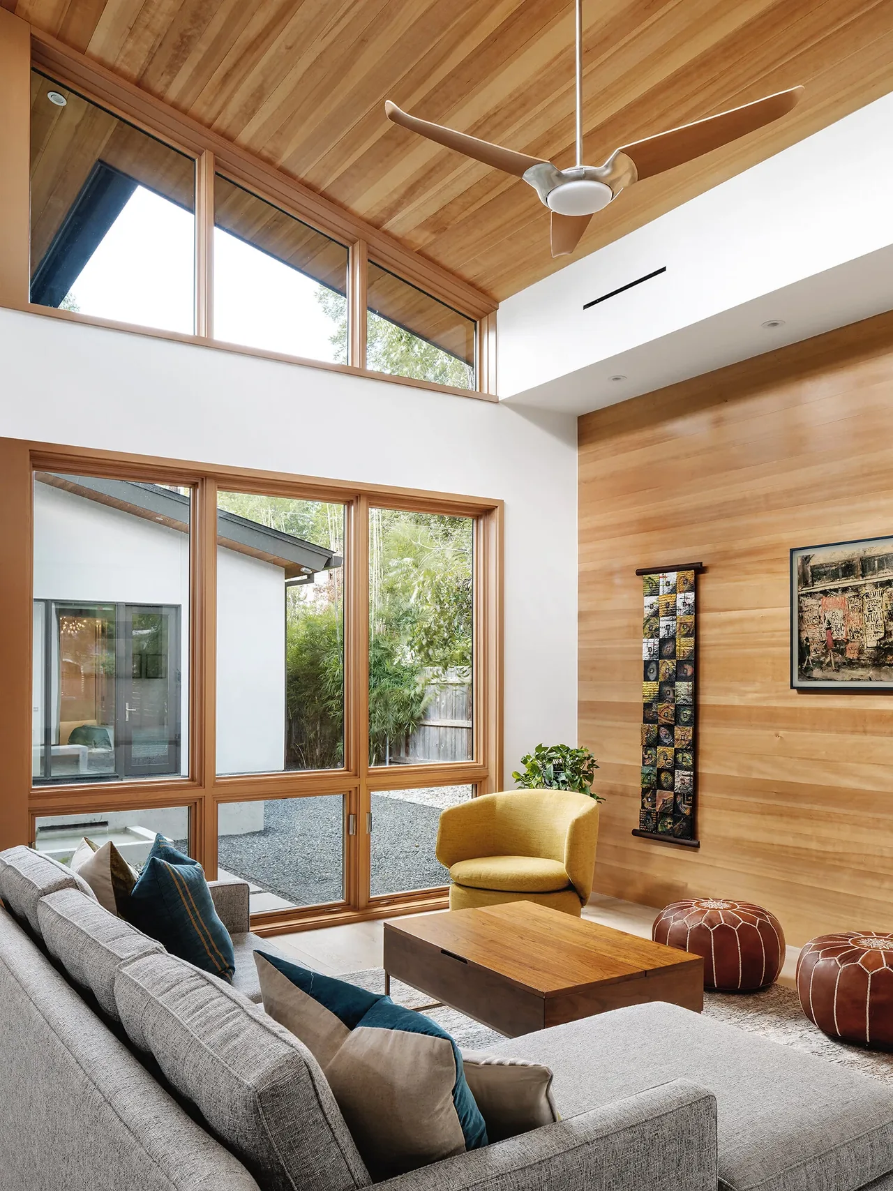 Living room of the Ramsey residence features a wood-paneled ceiling and wall, large wood-framed windows, and modern furnishings.