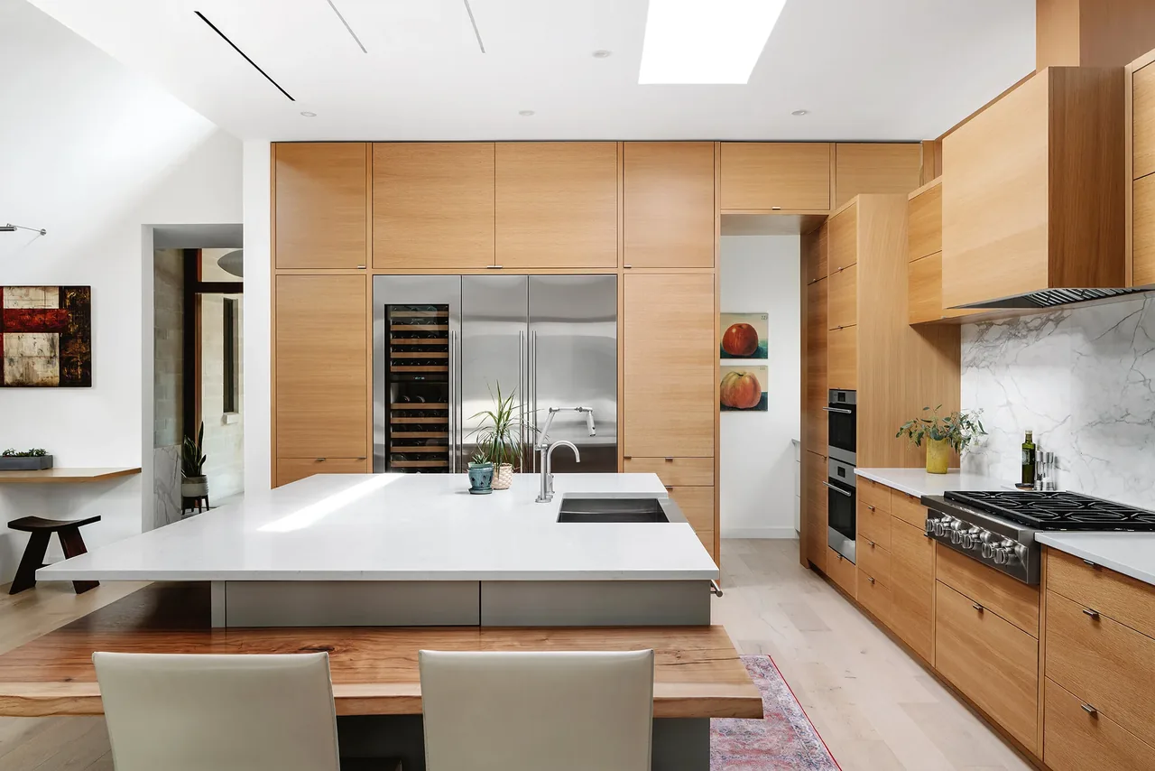 The Ramsey residence kitchen features a large white island with wood cabinetry and stainless steel appliances under skylights.