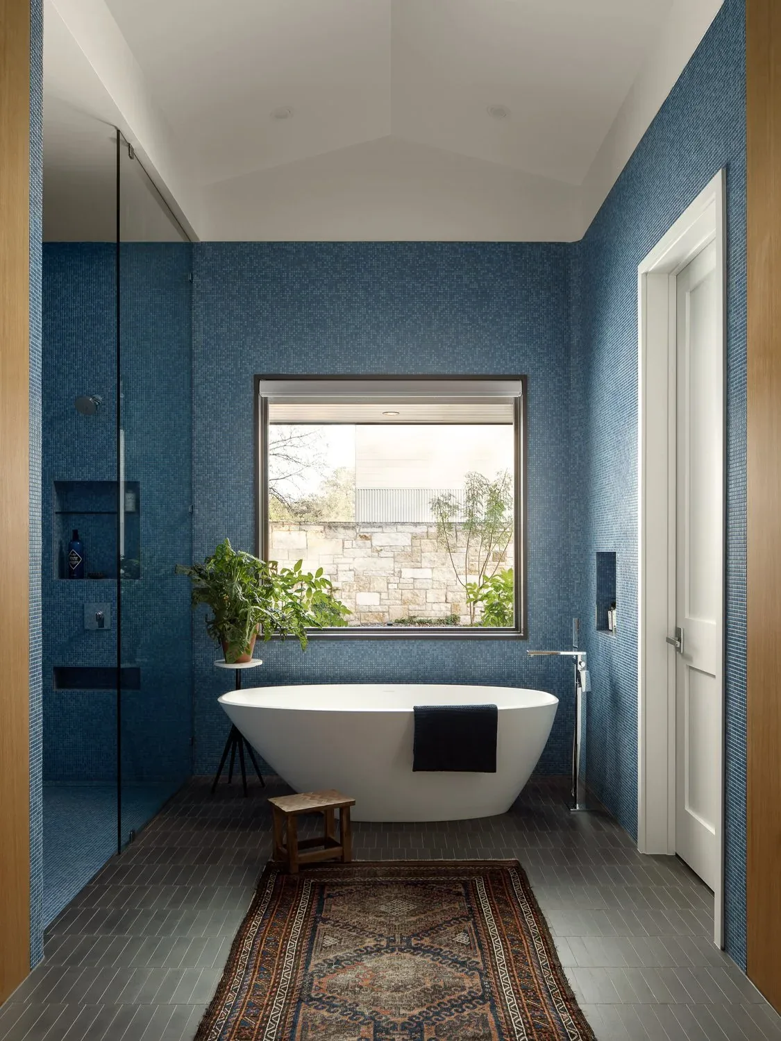 Freestanding tub bathroom with blue mosaic tile walls and a large window overlooking an outdoor stone wall.