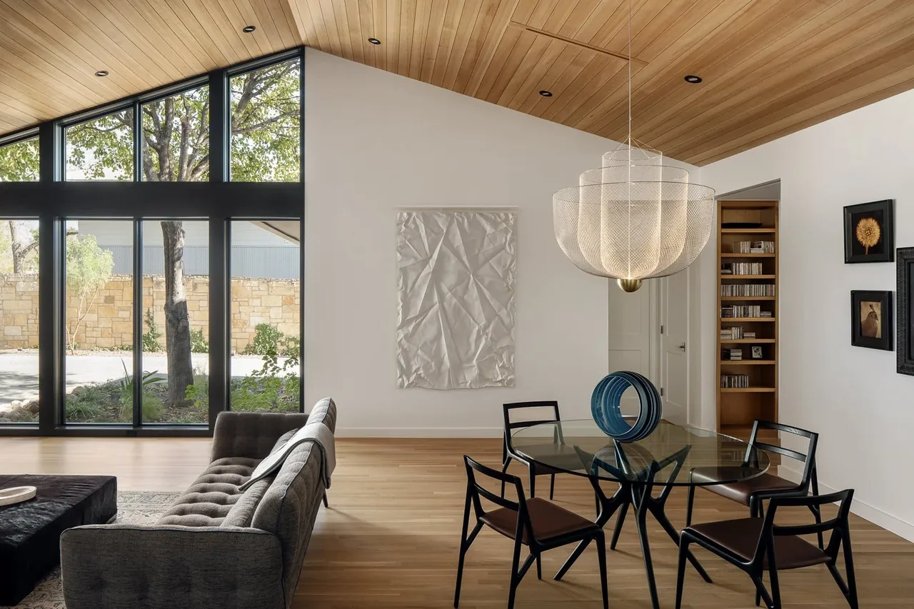 Dining space with a glass table and distinctive pendant light under a wood-paneled sloped ceiling, next to a living room with floor-to-ceiling windows.