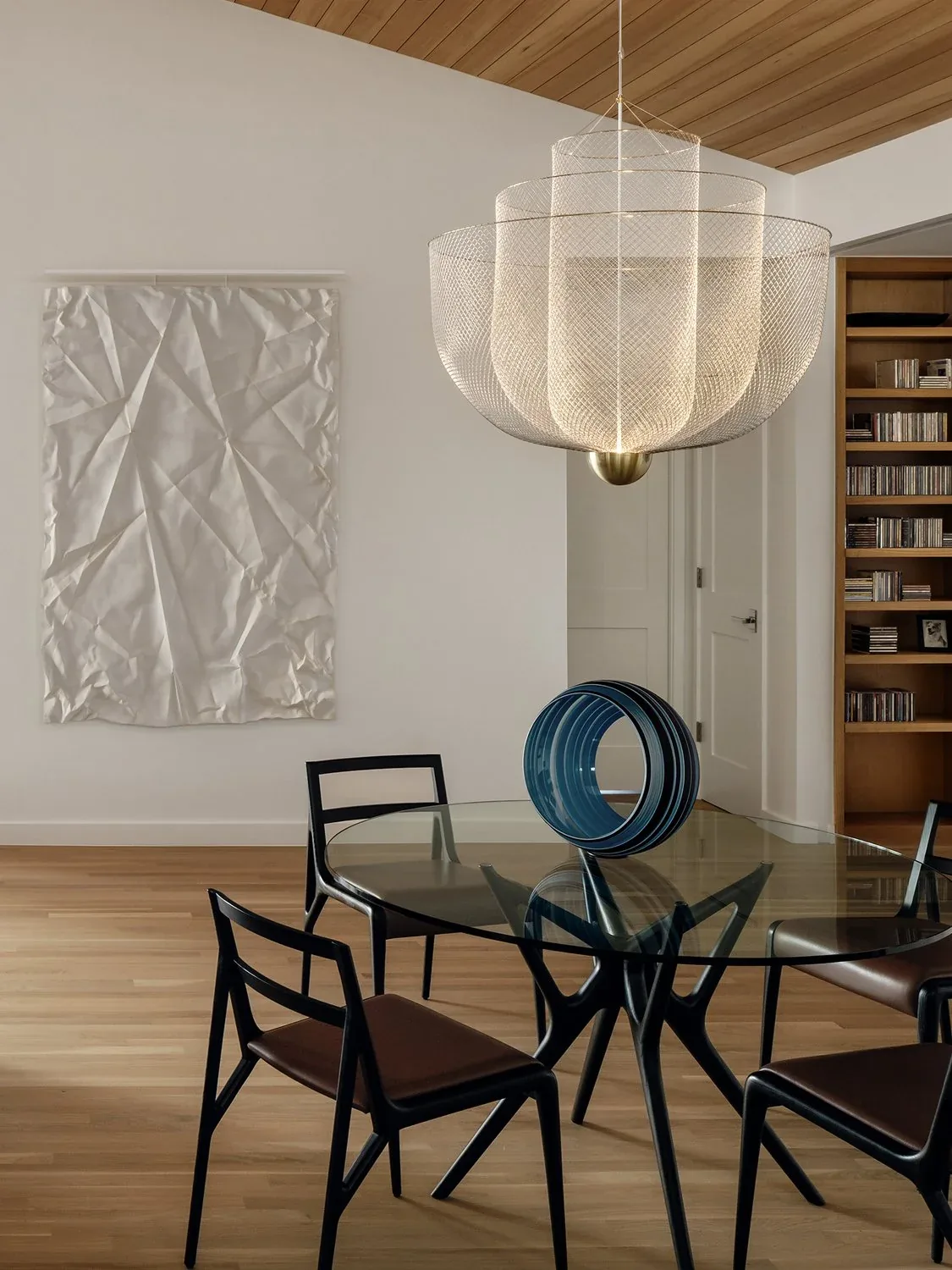 Dining space with a glass table and black chairs beneath a layered metal pendant light and wood-paneled ceiling.