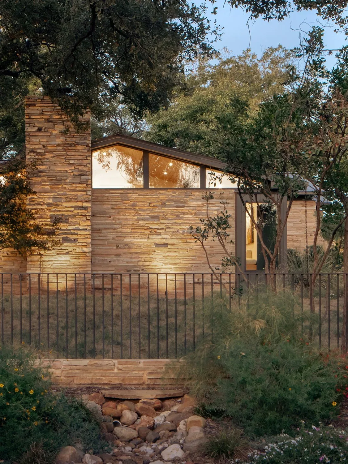 Stone-clad residence with clerestory windows and chimney seen in wooded setting with metal fence in foreground.