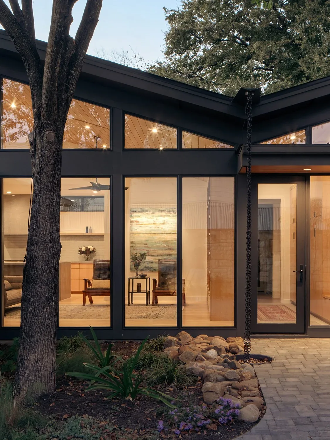 Modern residence exterior with black window frames, sloped clerestory windows, and a rain chain gutter over a rock bed.